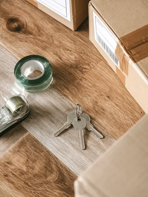 Top-down view of new apartment keys, a tape dispenser and labeled moving boxes on a wood floor