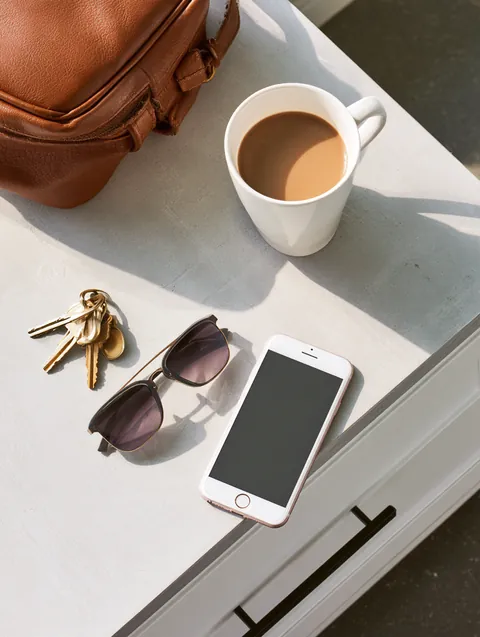 Top-down flat lay of house keys, sunglasses, a face-down smartphone and a coffee cup on an entryway console table