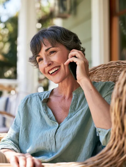 Woman in her fifties smiling with relief on her front porch while she talks on her smartphone about an emergency loan request through Hidden Meadow Lending